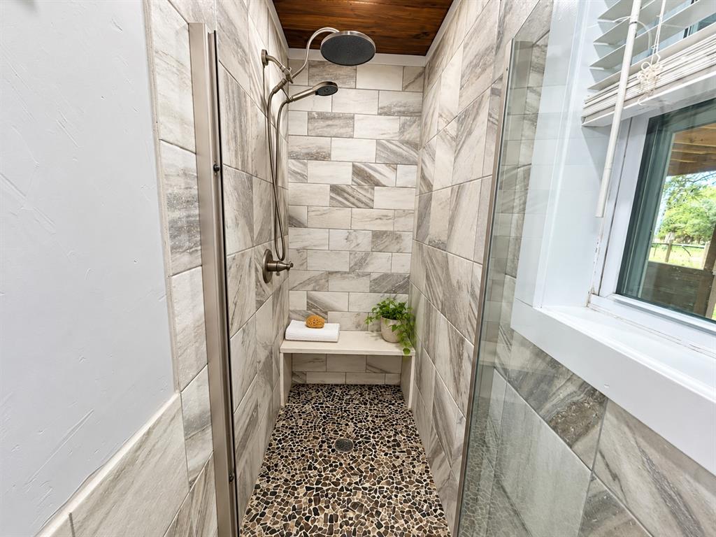 Walk-in shower featuring large-format neutral tile wall cladding, a built-in bench seat, a pebble-stone shower floor, a wood-plank ceiling, and a metallic dual-head shower fixture with glass surround