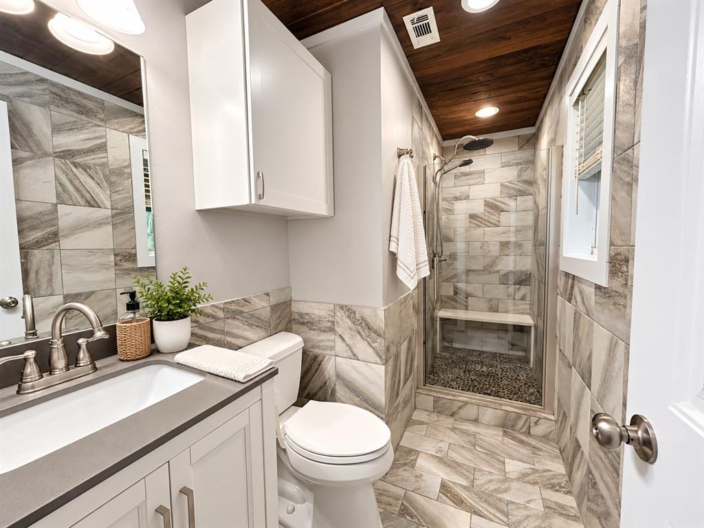 Bathroom featuring large-format decorative wall and floor tile, wood-plank ceiling, glass-enclosed shower with a built-in bench, and a solid-surface vanity countertop with an undermount sink