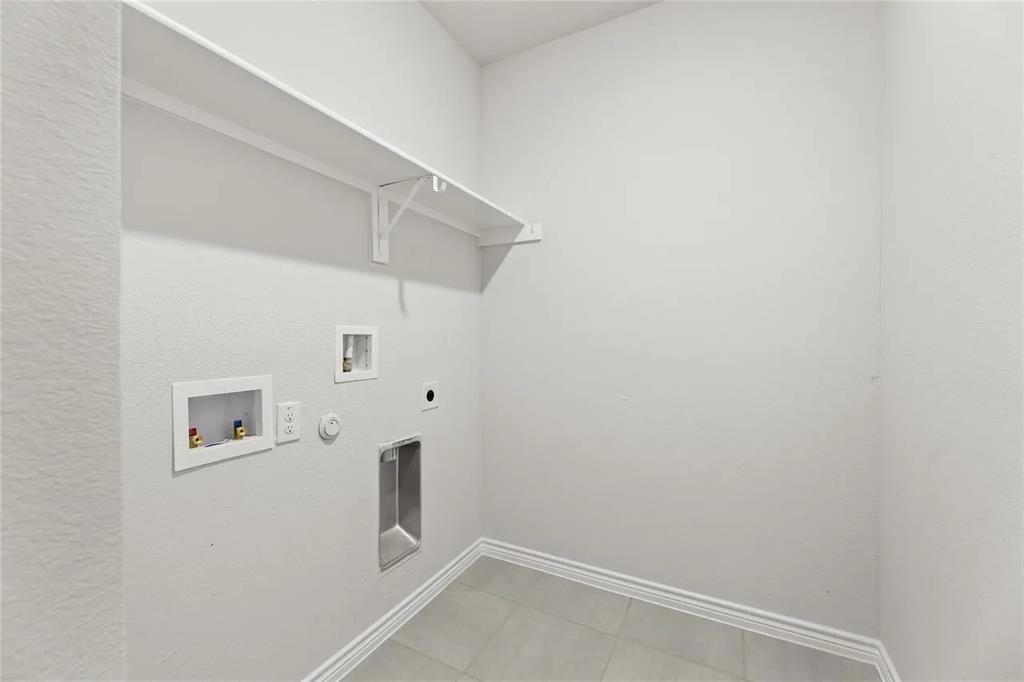 Utility area featuring neutral tile flooring, white wall-mounted shelving, and recessed plumbing connections