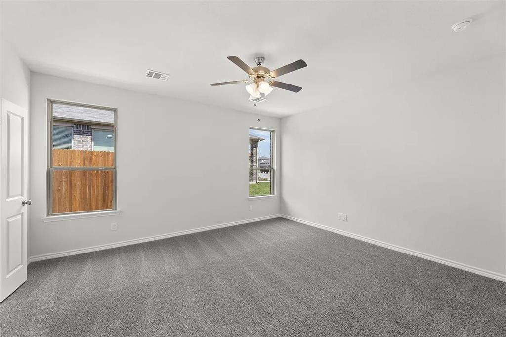 Neutral-toned interior room featuring wall-to-wall carpeting, dual windows with exterior views, and a multi-blade ceiling fan with integrated lighting