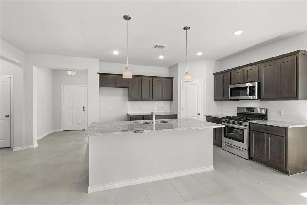 Kitchen featuring dark-toned cabinetry, stone-finish countertops, stainless steel appliances, a central island with an integrated sink, and dual overhead pendant lighting