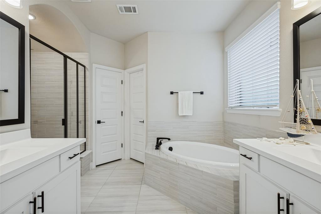 Bathroom featuring dual white vanities with solid-surface countertops, soaking tub with tile surround, glass-enclosed walk-in shower, and large-format tile flooring