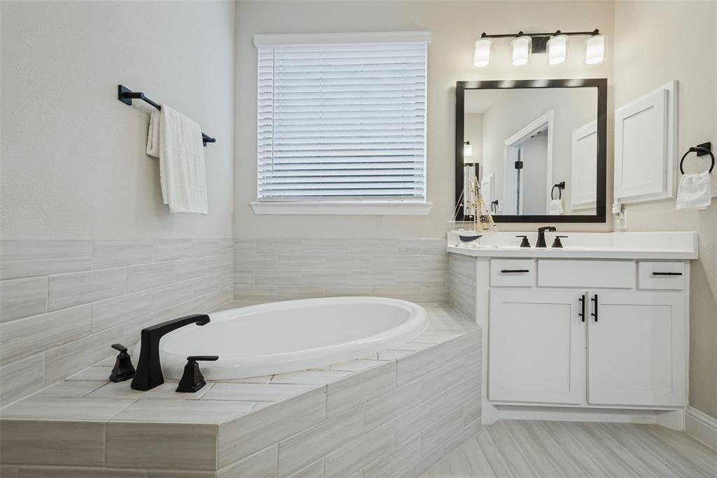 Soaking tub surrounded by horizontal tile, white vanity cabinet with a solid-surface countertop, matte black fixtures, framed vanity mirror, and linear vanity lighting