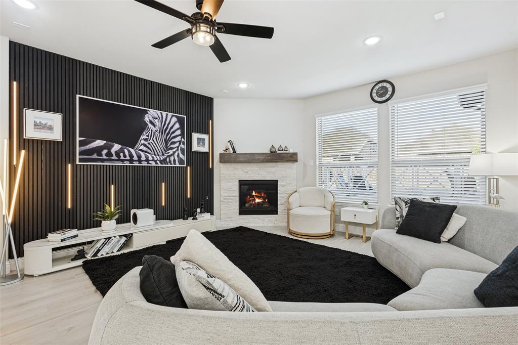 Living area featuring a vertical slat accent wall with integrated lighting, a stone-surround fireplace with a wooden mantel, wood-finish flooring, and a ceiling fan