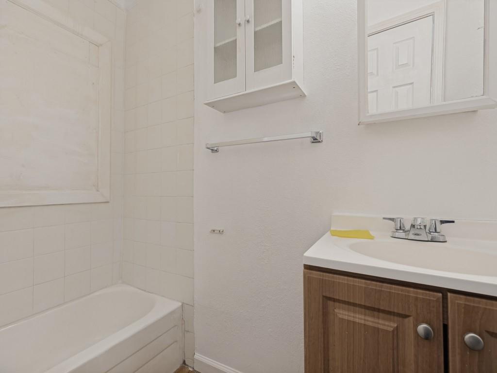 Bathroom featuring a wood-finish vanity with integrated sink, white tile tub surround, wall-mounted cabinet with glass doors, and a medicine cabinet with mirror