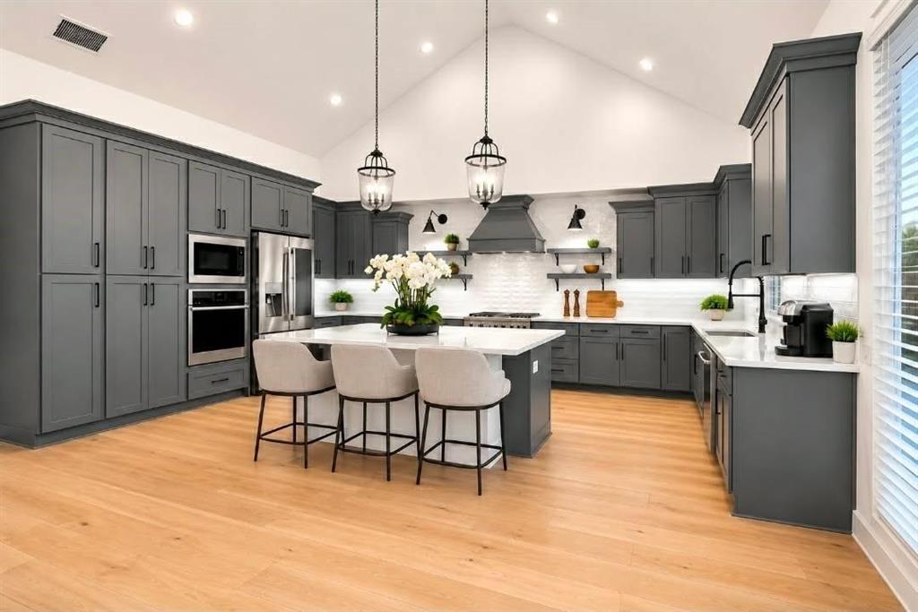 Kitchen featuring dark grey cabinetry, a central island with a stone-finish countertop, wood-finish flooring, a vaulted ceiling, and stainless steel built-in appliances Kitchen featuring dark grey cabinetry, a central island with a stone-finish countertop, wood-finish flooring, a vaulted ceiling, and stainless steel built-in appliances