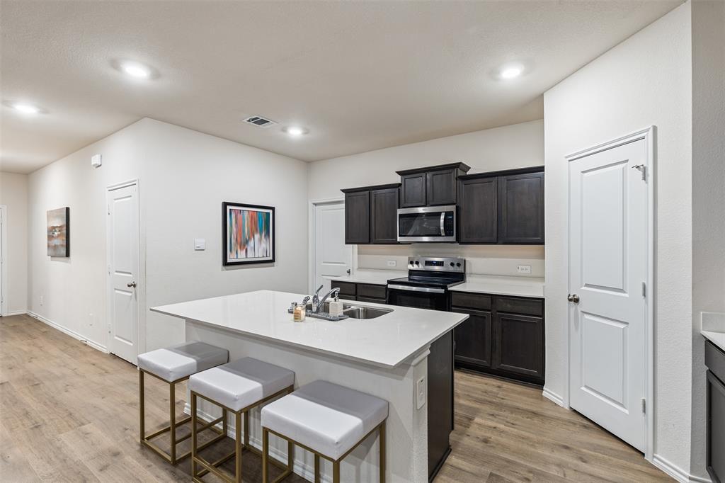 Kitchen featuring dark-finish cabinetry, white solid-surface countertops, a center island with an integrated stainless steel dual-basin sink, recessed ceiling lighting, and wood-finish flooring
