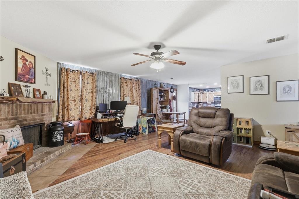 Brick fireplace with a rustic mantel, wood-finish flooring, metal accent wall, and a ceiling-mounted light fixture with a fan