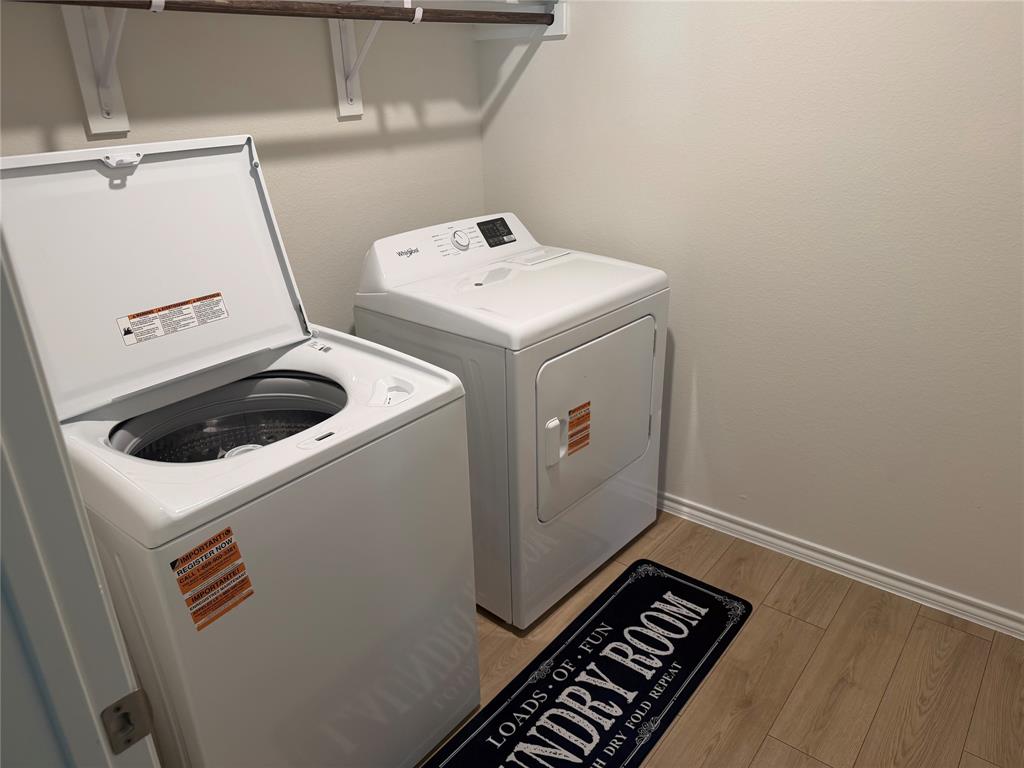 Laundry area featuring a top-load washer, front-load dryer, and wall-mounted shelving unit over wood-finish flooring Laundry area featuring a top-load washer, front-load dryer, and wall-mounted shelving unit over wood-finish flooring