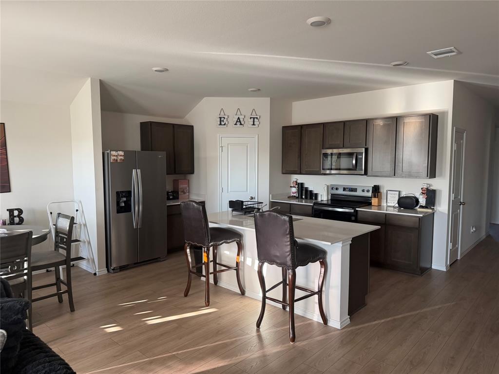 Kitchen featuring dark-finish cabinetry, stone-surface countertops, center island with seating, stainless steel appliances, and wood-finish flooring Kitchen featuring dark-finish cabinetry, stone-surface countertops, center island with seating, stainless steel appliances, and wood-finish flooring