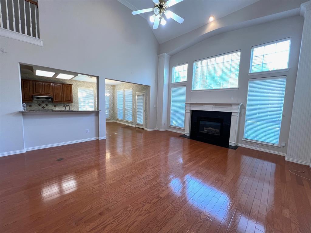 Vaulted ceiling with a central fan, hardwood flooring, fireplace with a tiered mantel and tile surround, oversized windows with blinds, and kitchen pass-through with a breakfast bar