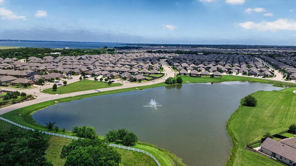Aerial perspective of a community pond featuring a central water fountain, surrounding green space, and white perimeter fencing