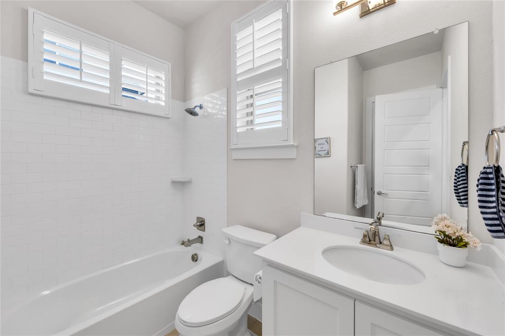 Bathroom featuring white cabinetry with a solid surface countertop, an undermount oval sink, shower-tub combination with white subway tile surround, plantation shutters, and a large wall-mounted mirror