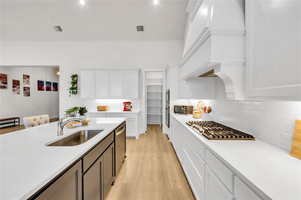 Kitchen featuring an island with a stainless steel undermount sink, white cabinetry, a built-in range hood, a tiled backsplash, and wood-finish flooring