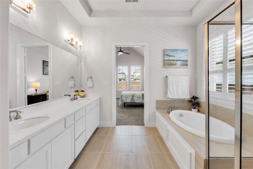 Dual-sink vanity with white cabinetry and solid-surface countertop, tile flooring, oval soaking tub, and glass-enclosed shower with plantation shutters