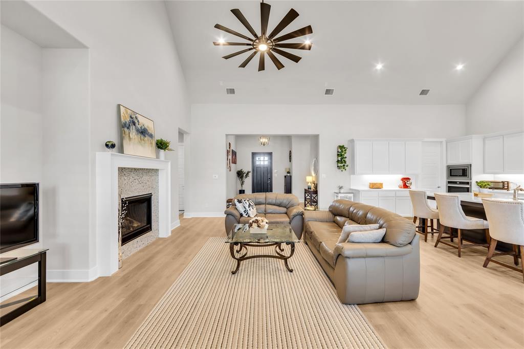 Vaulted ceiling featuring a central multi-blade light fixture, wood-finish flooring, stone-tiled fireplace, and kitchen with white cabinetry and a center island