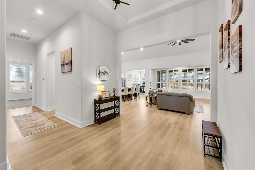 Open-concept interior featuring light wood-finish flooring, high ceilings with tray detailing, recessed lighting, and large windows with plantation shutters