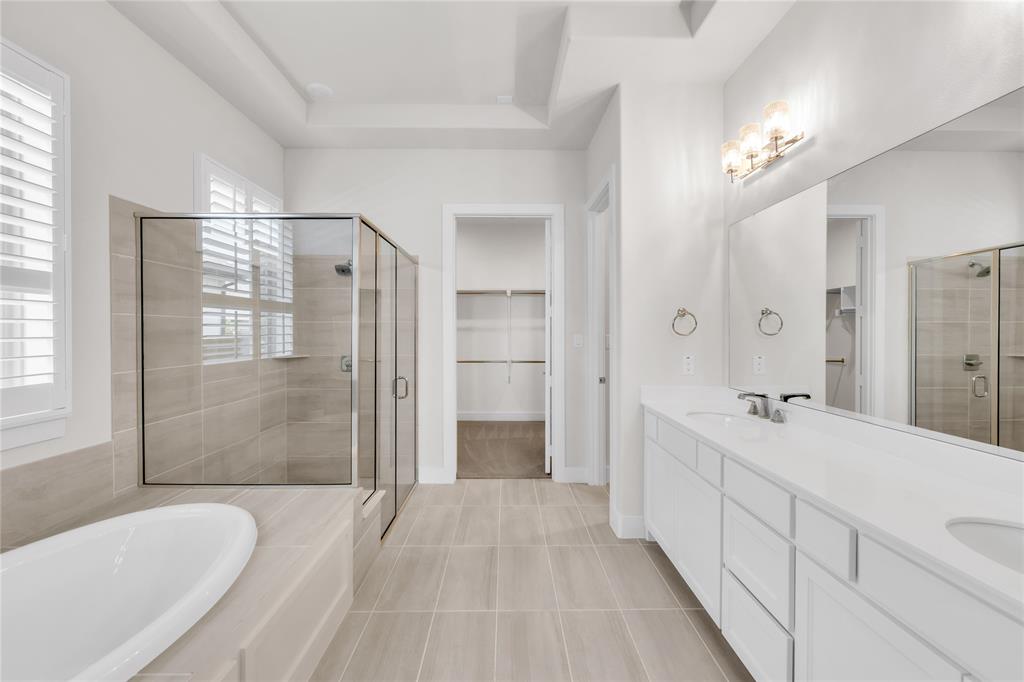 Dual-sink vanity with white cabinetry and solid-surface countertops, walk-in tile shower, separate soaking tub, and walk-in closet with built-in shelving