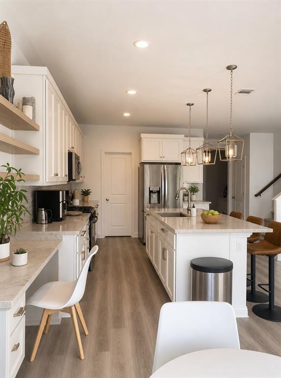 Kitchen featuring white cabinetry, wood-finish flooring, stone-composite countertops, a central island with an undermount sink, and three geometric pendant lights Kitchen featuring white cabinetry, wood-finish flooring, stone-composite countertops, a central island with an undermount sink, and three geometric pendant lights
