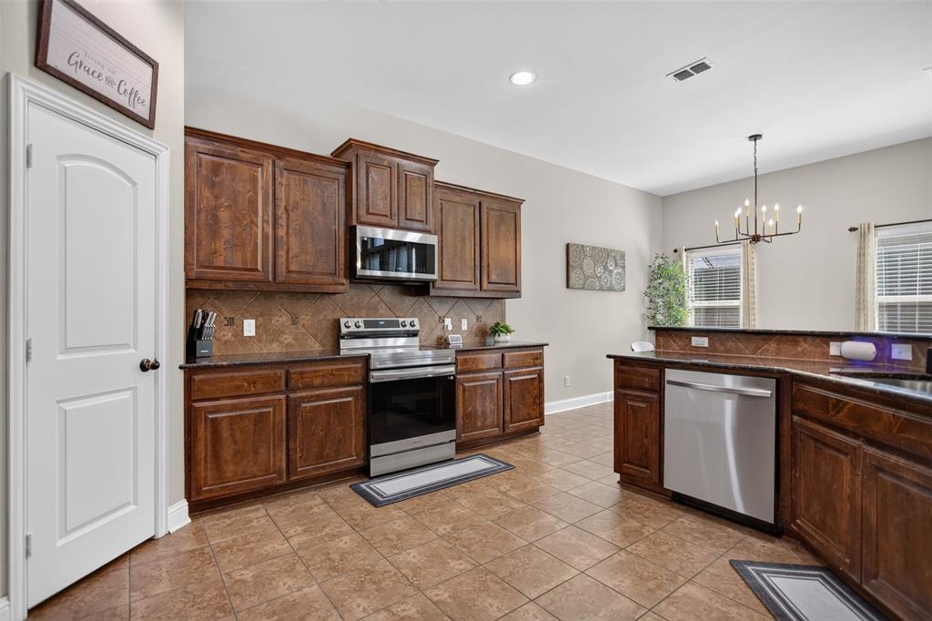 Kitchen featuring stained wood cabinetry, stone-tile backsplash, stainless steel appliances, dark countertops, and tile flooring