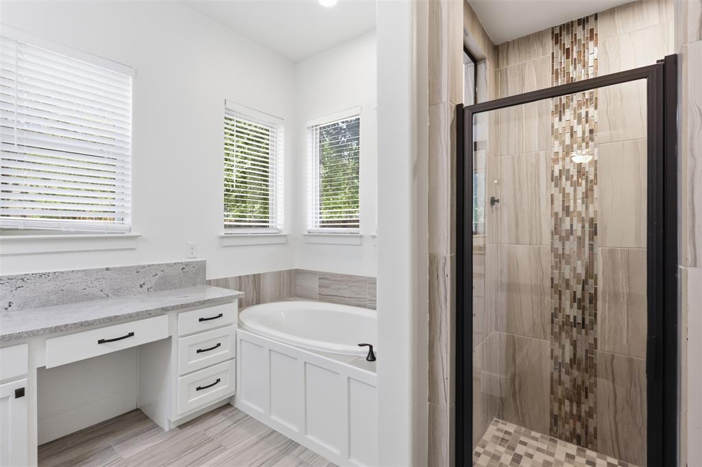 Bathroom featuring a built-in vanity with light-toned stone countertop, soaking tub with paneled surround, walk-in shower with decorative mosaic tile accent, and tile flooring