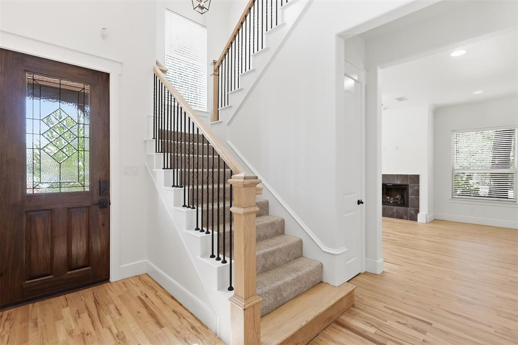 Stained wood entry door with decorative glass inlay, wood-finish flooring, staircase featuring a natural wood handrail and black metal balusters, and a fireplace with tile surround
