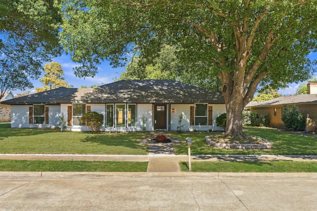 Single-story brick exterior featuring a low-pitched roof, wood shutter accents, front entry door with glass pane, and a mature tree within a stone-bordered landscape bed