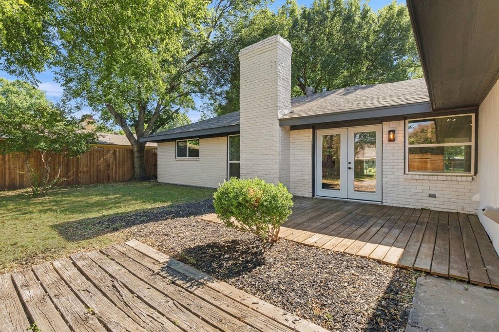 Exterior featuring white brick facade, masonry chimney, wood-plank decking, double glass-panel doors, and fenced backyard with mature trees