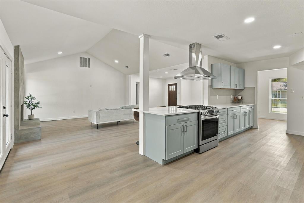 Vaulted ceilings, wood-finish flooring, kitchen island with stainless steel gas range, stainless steel vent hood, and light-toned shaker-style cabinetry
