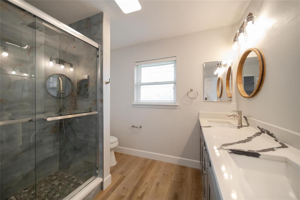 Bathroom featuring a walk-in shower with stone tile surround, double-sink vanity with patterned stone countertop, circular wood-frame mirrors, and wood-finish flooring