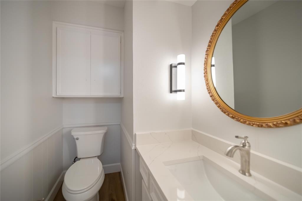 Vanity with quartz countertop and integrated sink, wall-mounted light fixture, oval mirror with ornate metallic frame, white beadboard wainscoting, and built-in upper cabinet storage
