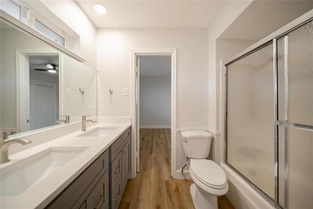 Dual-sink vanity with white countertops and gray cabinetry, large wall-mounted mirror, wood-finish flooring, and a shower-tub combination with sliding glass doors