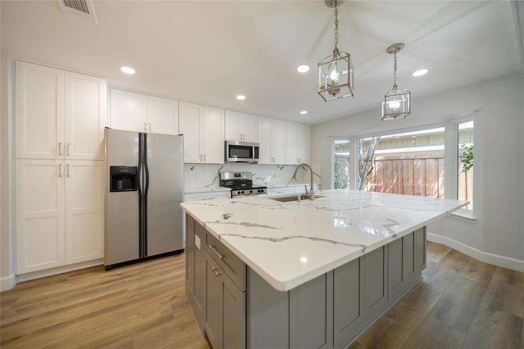 Kitchen featuring white cabinetry, an expansive center island with stone-finish countertops, stainless steel appliances, wood-finish flooring, and dual lantern-style pendant lights Kitchen featuring white cabinetry, an expansive center island with stone-finish countertops, stainless steel appliances, wood-finish flooring, and dual lantern-style pendant lights