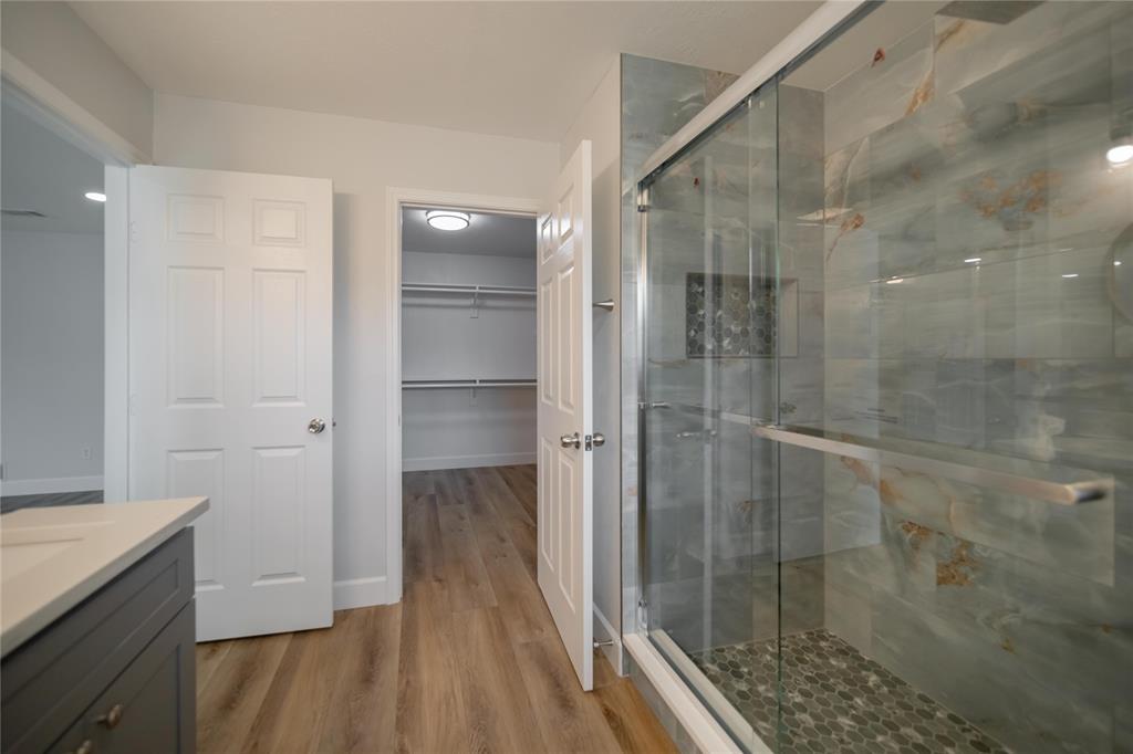 Walk-in shower featuring large-format tile walls, integrated shelving niche, and pebble-style floor tiling, complemented by a wood-finish vanity and walk-in closet with built-in shelving