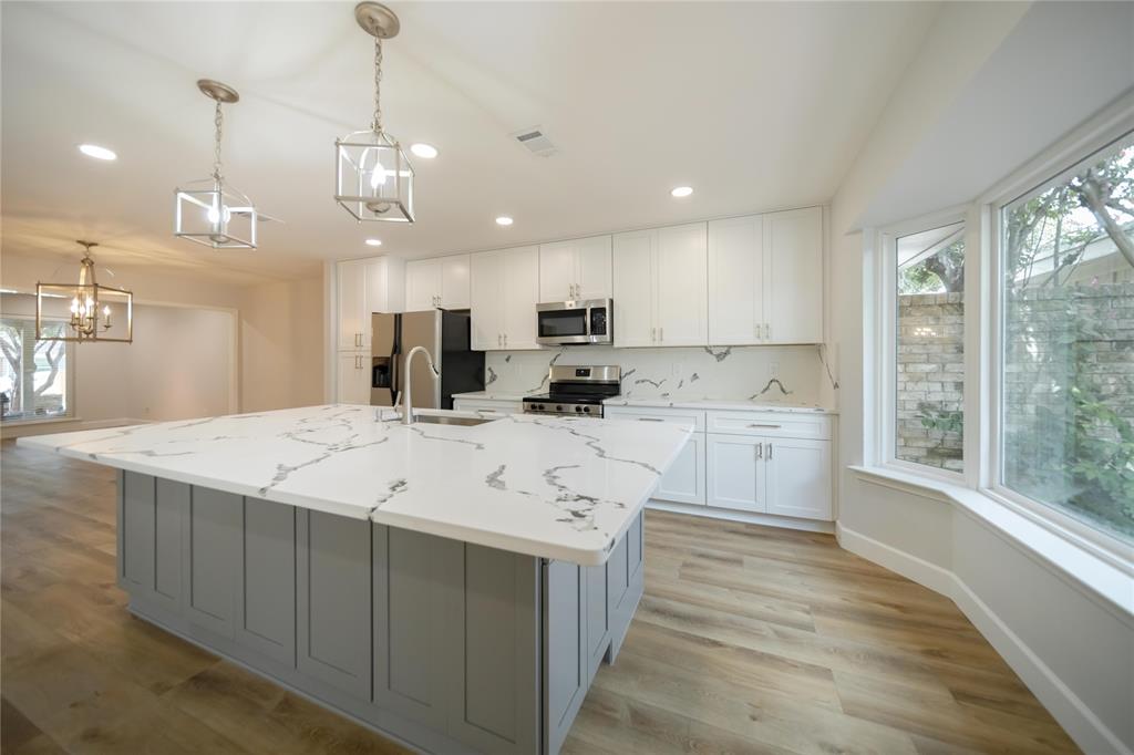 Kitchen featuring an oversized central island with stone-finish countertops, white cabinetry, stainless steel appliances, pendant lighting, and wood-finish flooring Kitchen featuring an oversized central island with stone-finish countertops, white cabinetry, stainless steel appliances, pendant lighting, and wood-finish flooring