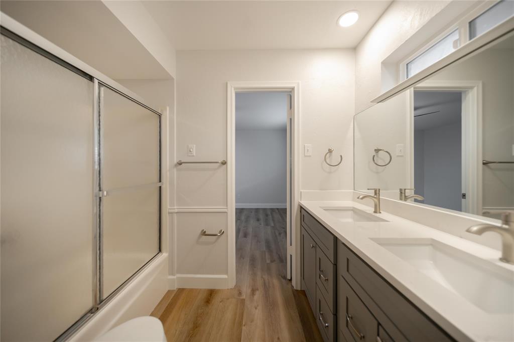 Bathroom featuring a double vanity with grey cabinetry, a solid surface countertop with integrated dual sinks, a full-width vanity mirror, wood-finish flooring, and a tub-shower combination with a sliding glass enclosure