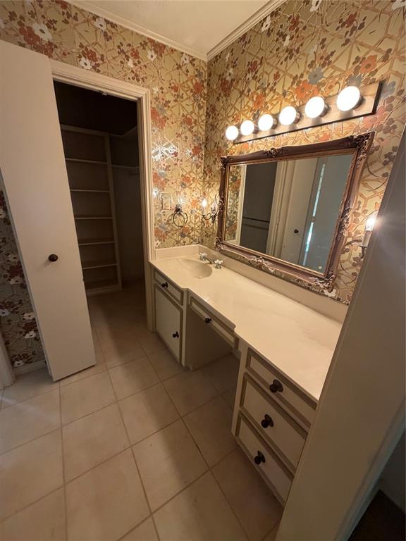 Built-in vanity featuring a solid surface countertop, integrated sink, and ample cabinetry, paired with a framed wall mirror and multi-bulb vanity light, finished with large-format tile flooring and a walk-in storage area with built-in shelving