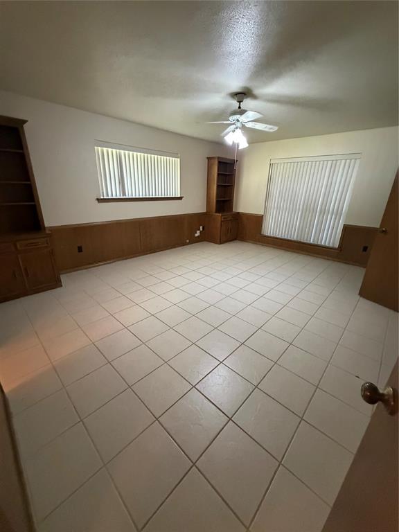 Room featuring ceramic tile flooring, wood paneling, dual built-in wooden shelving units, and a ceiling fan with integrated light fixture Room featuring ceramic tile flooring, wood paneling, dual built-in wooden shelving units, and a ceiling fan with integrated light fixture