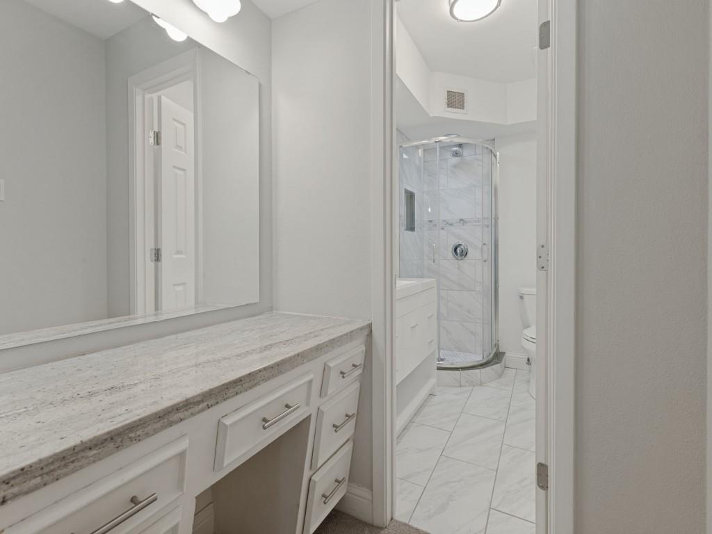 White vanity with stone countertop, large wall-mounted mirror, corner shower enclosure with tiled surround, and tile flooring