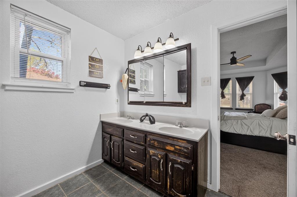 Dual-sink vanity with dark wood cabinetry and a solid-surface countertop, illuminated by a four-light fixture above a tri-fold medicine cabinet, complemented by large-format tile flooring