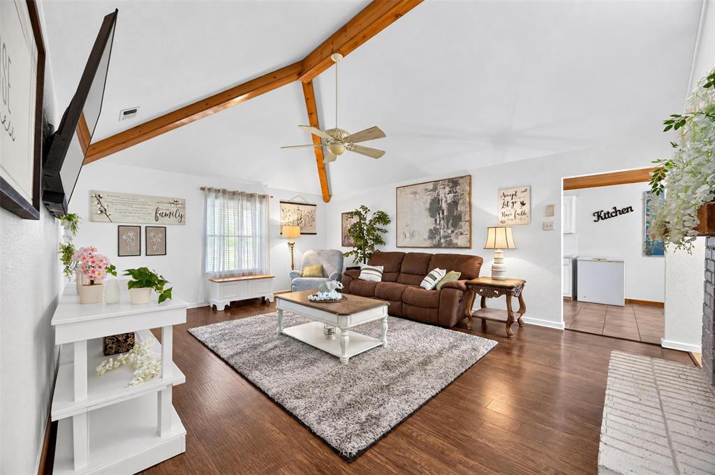 Vaulted ceiling with exposed wood beams, wood-finish flooring, ceiling fan, and brick fireplace surround