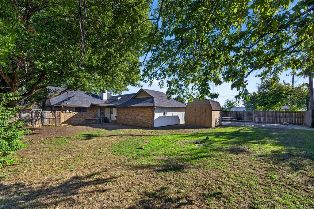 Spacious backyard featuring brick exterior siding, a shingled roof, mature tree canopy, and an attached storage structure enclosed by perimeter wood fencing