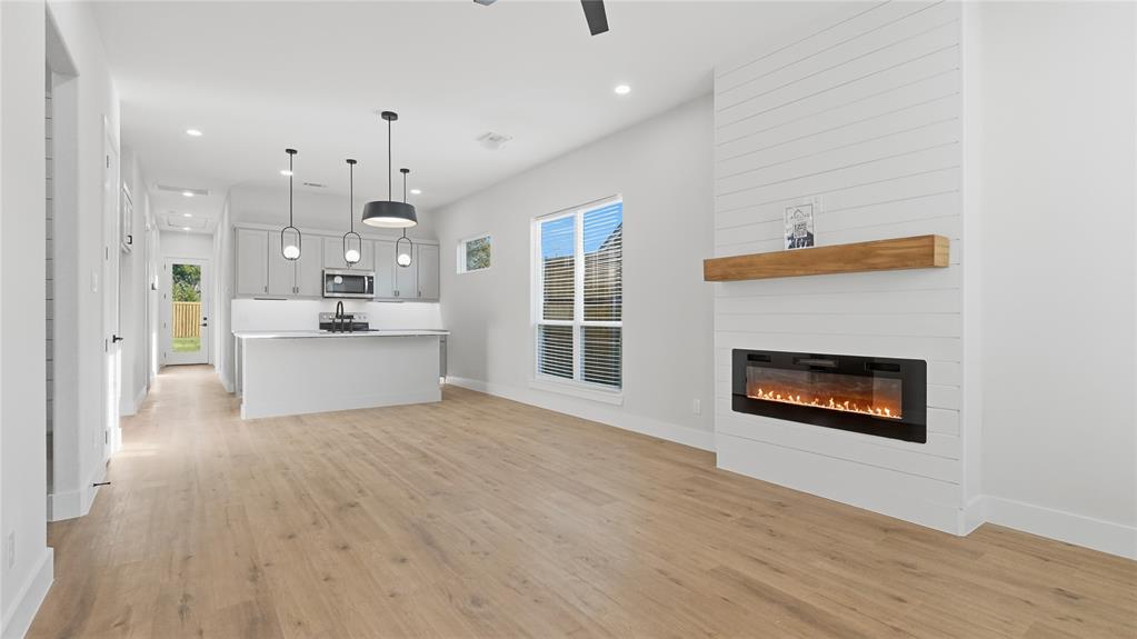 Open-concept area featuring wood-finish flooring, a white shiplap fireplace surround with a natural wood mantel, an electric wall-mounted fireplace, a kitchen island with pendant lighting, and recessed ceiling lighting