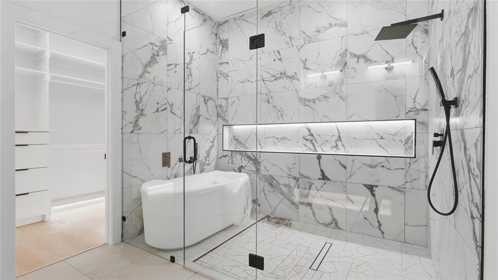 Wet room featuring a freestanding soaking tub, large-format tile surround, rain shower head, handheld sprayer, and recessed wall niche with integrated lighting, enclosed by a frameless glass partition