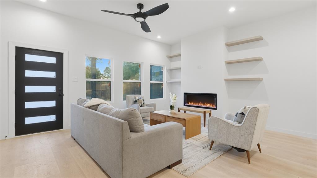 Modern interior featuring light-toned wood-finish flooring, a linear electric fireplace, recessed lighting, matte black ceiling fan, and built-in floating shelves