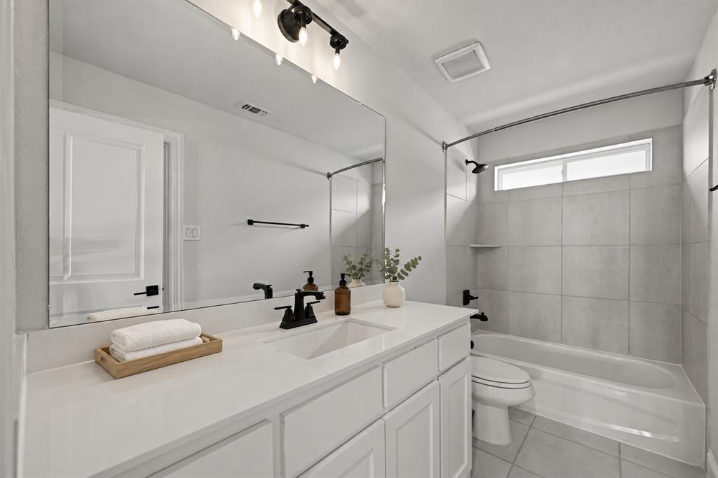 White vanity cabinetry with a stone countertop, integrated sink, large wall-mounted mirror, matte black plumbing fixtures, and a tub-shower combination featuring tiled wall surround
