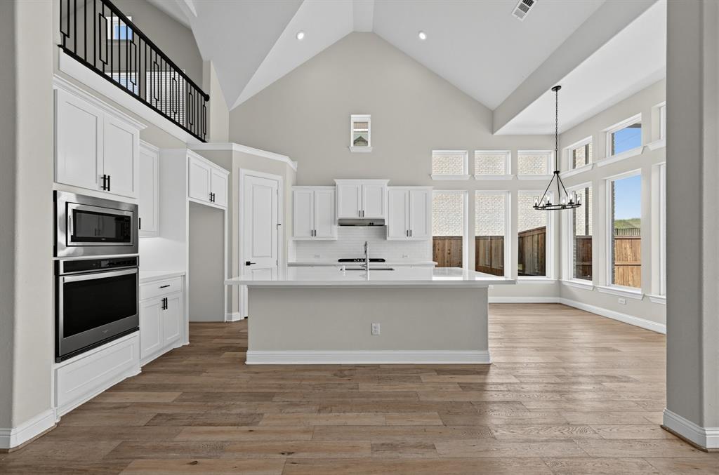 Vaulted ceiling with an elevated mezzanine featuring a black metal railing, expansive windows, center island with stone-finish countertop, wood-finish flooring, and built-in stainless steel double wall ovens