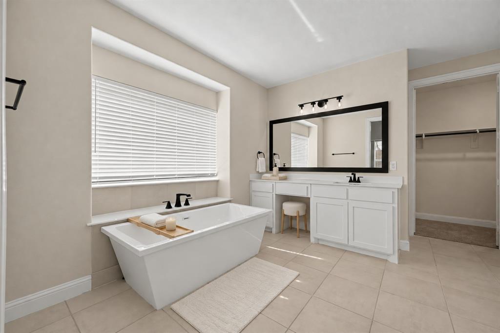 Freestanding soaking tub with matte black deck-mounted faucet, white cabinetry with integrated vanity seating, large rectangular mirror with black frame, tile flooring, and light-filtering horizontal blinds