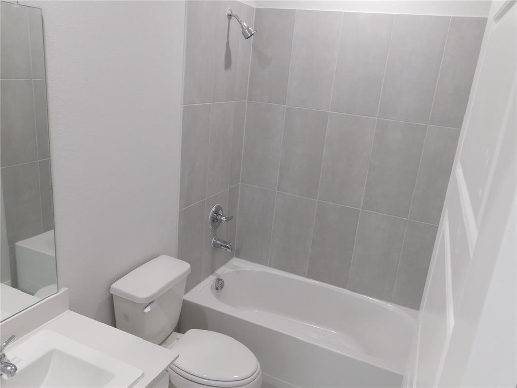 Bathroom featuring a porcelain tub and shower combination with full-height grey tile surround, white solid-surface vanity countertop, undermount sink with chrome hardware, and wall-mounted vanity mirror