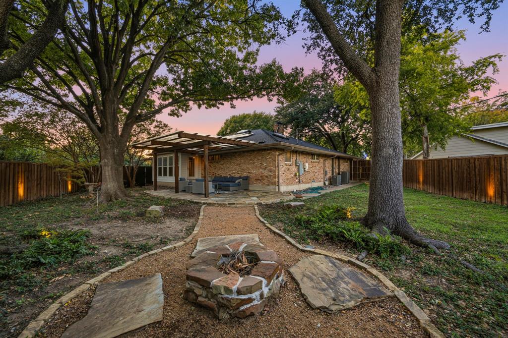 Backyard featuring a stone-clad fire pit, a covered pergola, a natural stone path, mature shade trees, and perimeter wood fencing with integrated accent lighting
